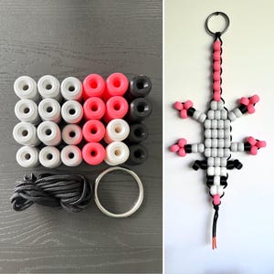 May include: A craft kit containing white, neon pink, and black pony beads, a length of black cord, and a metal ring, alongside a finished lizard-shaped keychain assembled from the same beads, cord, and ring, featuring a long tail and four legs.