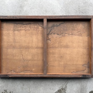 May include: A rectangular wooden tray with a dark brown frame and a light brown interior. The tray is divided into two sections by a vertical divider. The wood grain is visible, and there are some dark stains on the surface.