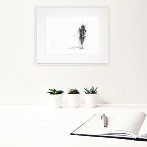 May include: A black and white sketch of a person walking, framed in a white frame and hanging on a white wall. Three small potted plants are on a white surface in front of the frame.
