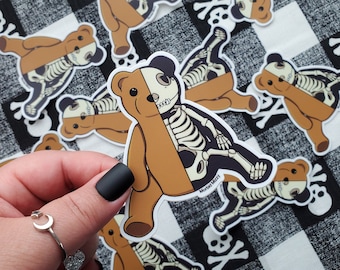 May include: A set of brown teddy bear stickers with a skeleton inside. The stickers are on a black and white checkered background. The text on the sticker says "Mortal Mazzi".
