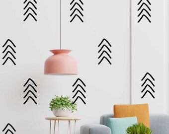 May include: A white wall with black geometric shapes in the form of arrows. The shapes are arranged in a symmetrical pattern. A pink lamp hangs from the ceiling. A small potted plant sits on a metal table. A gray couch with a yellow and blue pillow sits in the foreground.