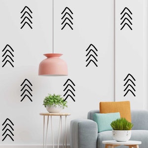 May include: A white wall with black geometric shapes in the form of arrows. The shapes are arranged in a symmetrical pattern. A pink lamp hangs from the ceiling. A small potted plant sits on a metal table. A grey sofa with a yellow and blue cushion sits in the foreground.