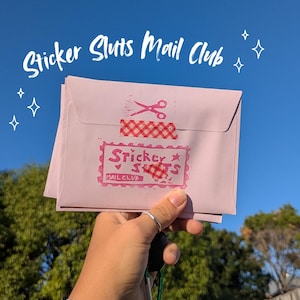 K&ouml;nnte beinhalten: Ein hellrosa Umschlag mit einem rot-wei&szlig; karierten Klebebandverschluss, einem gestempelten rosa Scherensymbol und einem rechteckigen rosa Stempel mit dem Text Sticker Sluts Mail Club. Der Text Sticker Sluts Mail Club steht &uuml;ber dem Umschlag.