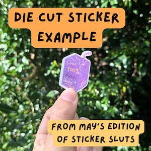 K&ouml;nnte beinhalten: Ein gestanzter Aufkleber in Form einer lila Saftpackung mit Strohhalm. Der Aufkleber hat ein holografisches Finish und den Text 100% delulu JUICE. Das Design enth&auml;lt kleine Stern- und Kringelmotive in einer helllila und rosafarbenen Palette.