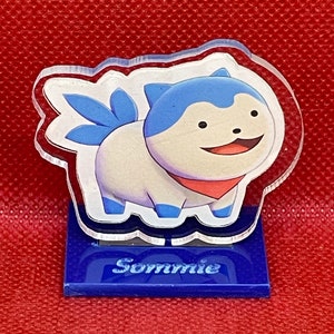 May include: A clear acrylic figure of a blue and white cartoon dog with a red bandana. The figure is standing on a blue base with the word "Sommie" printed on it.