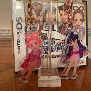 Könnte beinhalten: Zwei Schlüsselanhänger aus Acryl mit Anime-Charakteren, einer in Rosa mit Hasenohren, der andere in einem lila und rosa Kimono. Beide haben goldfarbene Verschlüsse und werden vor einer Nintendo DS-Spielhülle ausgestellt.