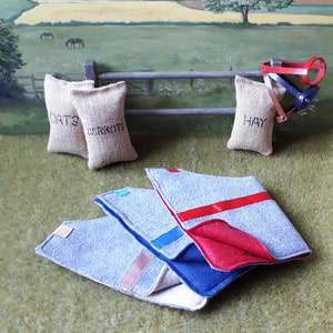 Puede incluir: Tres mantas de caballo de fieltro gris con ribetes rojos, azules y rosas. Las mantas est&aacute;n dobladas y apiladas sobre una superficie de hierba verde. Tres sacos de arpillera etiquetados con "Avena", "Zanahorias" y "Heno" est&aacute;n en el fondo.
