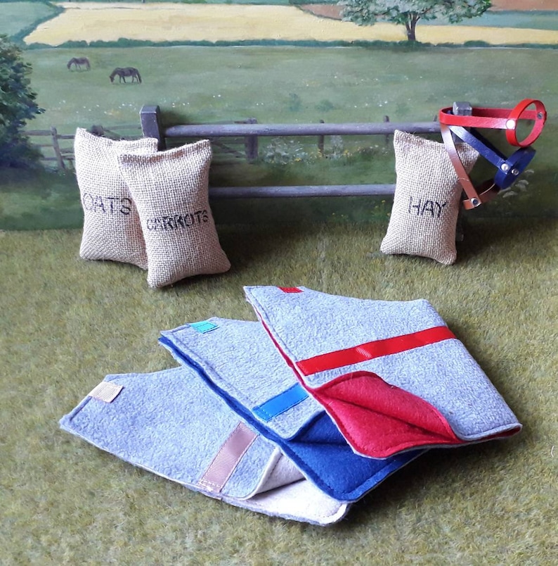 Puede incluir: Tres mantas de caballo de fieltro gris con ribetes rojos, azules y rosas. Las mantas est&aacute;n dobladas y apiladas sobre una superficie de hierba verde. Tres sacos de arpillera etiquetados con "Avena", "Zanahorias" y "Heno" est&aacute;n en el fondo.
