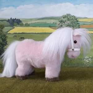 K&ouml;nnte beinhalten: Ein rosa Pl&uuml;schpony mit wei&szlig;er M&auml;hne und Schweif, das ein wei&szlig;es Lederhalfter tr&auml;gt. Das Pony steht auf einer gr&uuml;nen Grasfl&auml;che mit einem gemalten Hintergrund einer Landschaft.
