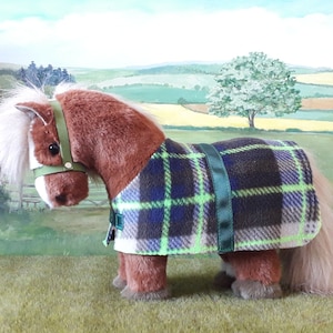 Puede incluir: Un caballo de peluche marr&oacute;n que lleva una manta de forro polar a cuadros verde, azul y blanco con una cabezada verde.