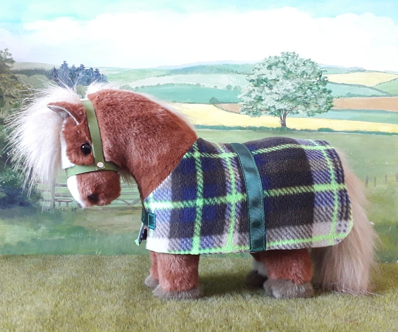 Puede incluir: Un caballo de peluche marr&oacute;n que lleva una manta de forro polar a cuadros verde, azul y blanco con una cabezada verde.