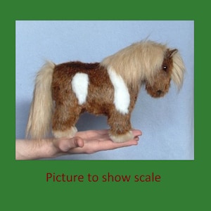 K&ouml;nnte beinhalten: Ein braunes und wei&szlig;es Pl&uuml;schpony mit einer langen M&auml;hne und einem langen Schwanz. Das Pony steht auf der Hand einer Person, um seine Gr&ouml;&szlig;e zu zeigen.