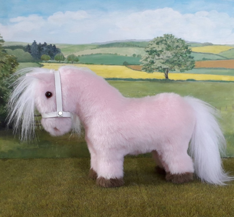 K&ouml;nnte beinhalten: Ein rosa Pl&uuml;schpony mit wei&szlig;er M&auml;hne und Schweif. Das Pony tr&auml;gt ein wei&szlig;es Lederhalfter.