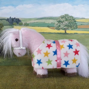 K&ouml;nnte beinhalten: Ein rosa Pl&uuml;schpony, das eine wei&szlig;e Fleece-Decke mit bunten Sternen tr&auml;gt. Die Decke hat einen rosa Schleifenbesatz und das Pony tr&auml;gt ein wei&szlig;es Halfter.