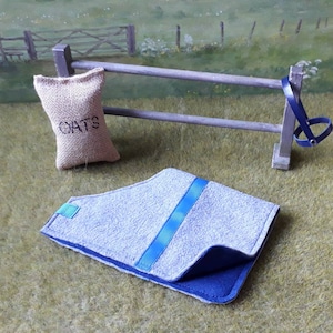 Puede incluir: Una manta de caballo gris y azul con una cinta azul en el centro. La manta est&aacute; doblada por la mitad y est&aacute; sobre una superficie de hierba verde. Una bolsa de arpillera etiquetada con "OATS" cuelga de una valla de madera en el fondo.