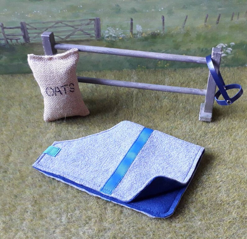 Puede incluir: Una manta de caballo gris y azul con una cinta azul en el centro. La manta est&aacute; doblada por la mitad y est&aacute; sobre una superficie de hierba verde. Una bolsa de arpillera etiquetada con "OATS" cuelga de una valla de madera en el fondo.