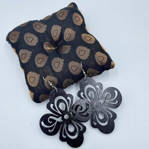 May include: A pair of black, laser-cut flower earrings with silver-colored hooks. The earrings are displayed on a small, patterned pillow with a black background and gold-colored design.