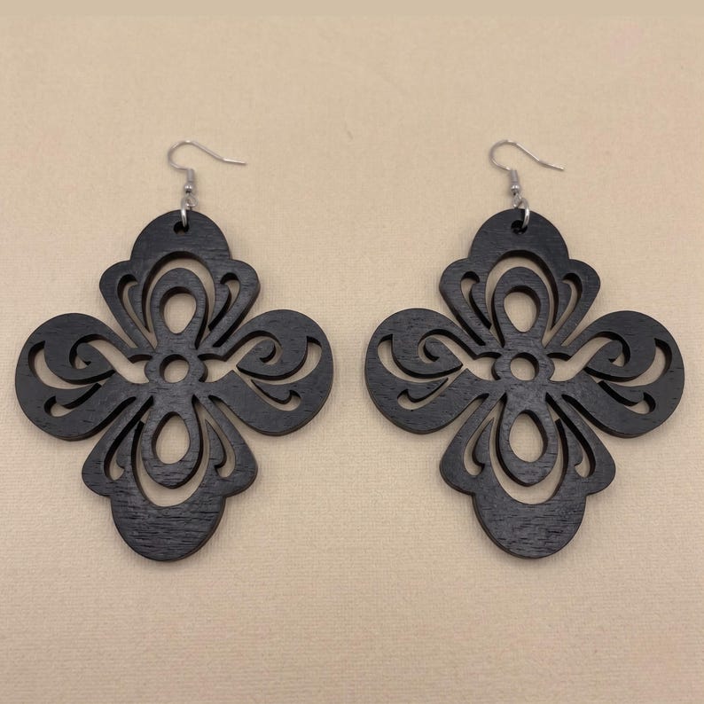 May include: A pair of black wooden earrings. Each earring features a symmetrical, openwork design with a floral or quatrefoil shape. The earrings have silver-colored fish hook ear wires.