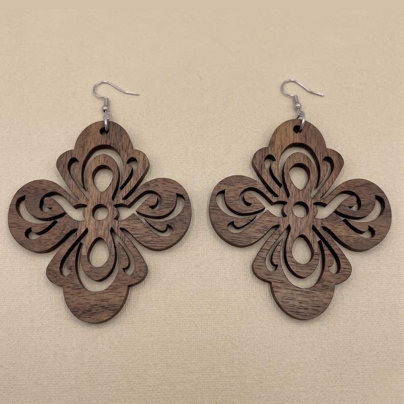 May include: A pair of wooden earrings with a dark brown finish. Each earring features a four-leaf clover design with intricate cut-out patterns and silver-colored ear wires. The earrings are displayed on a neutral background.