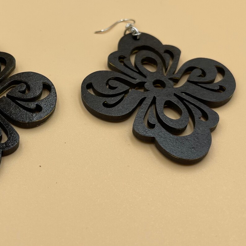 May include: Black, laser-cut wooden earrings with a floral design. The earrings have a four-petal shape with intricate cut-out details and silver-colored ear wires. The earrings are displayed against a neutral background.