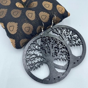 May include: Black wooden tree of life earrings. The earrings are circular with a detailed tree design cut out. The earrings hang from silver-colored hooks. The earrings are displayed on a black and gold patterned pillow.