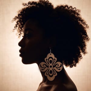 May include: Silhouette of a person with curly hair, wearing large, light-colored wooden earrings. The earrings have a decorative, openwork design. The background is a warm, neutral tone, creating a contrast with the dark hair and skin.