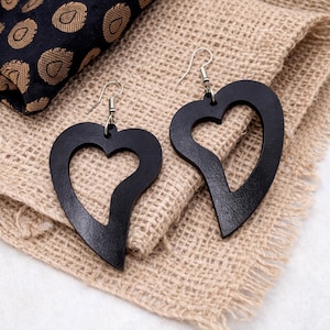 May include: A pair of black heart-shaped earrings with a cutout design. The earrings are made of a dark material and hang from silver-colored hooks. They rest on a textured, tan fabric, with a patterned dark blue and gold fabric in the background.