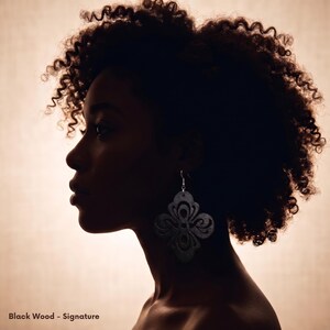 May include: Silhouette of a person with dark, curly hair wearing a large, black, floral-shaped earring. The text "Black Wood - Signature" is at the bottom of the image. The background is a warm, neutral tone.