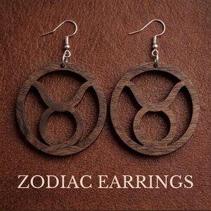 May include: A pair of circular dark brown wooden earrings featuring a cutout of the Taurus zodiac symbol. Each earring is suspended from a silver-toned metal hook. The text ZODIAC EARRINGS is printed in white at the bottom of the image.
