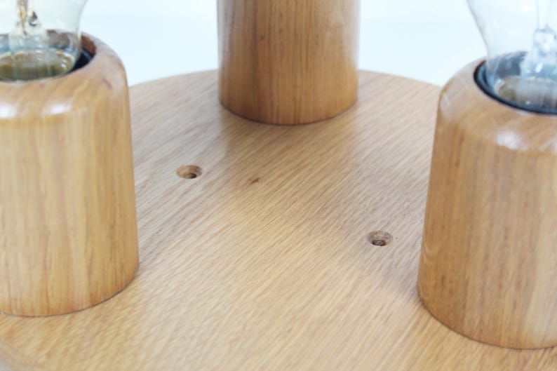 May include: Close-up of a wooden light fixture base with three cylindrical bulb holders. The base is a light-colored wood, and the holders are made of the same material. The fixture is designed to hold light bulbs.