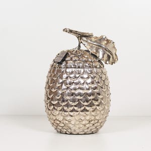 Op de afbeelding: Een decoratieve houder in de vorm van een ananas met een zilverkleurige, schubachtige textuur. Het object is voorzien van een opvallend, gevormd blad op het deksel en bevat een klein, rechthoekig zwart label dat eraan vastzit.