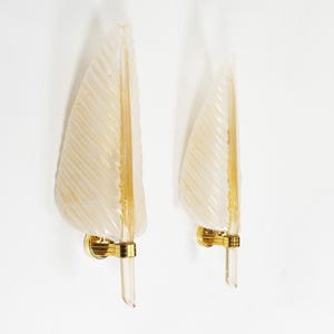 May include: A pair of wall sconces featuring curved, ribbed, off-white glass shades shaped like leaves. Each light fixture is supported by a polished gold-tone metal mount and a vertical glass rod, creating a sleek, vertical design for wall lighting.