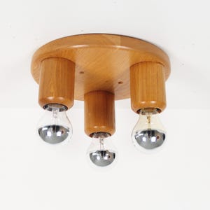 May include: A wooden ceiling light fixture with three cylindrical light holders. The fixture has a round, light-brown wooden base. Each holder has a silver-colored bulb. The light fixture is mounted on a white ceiling.