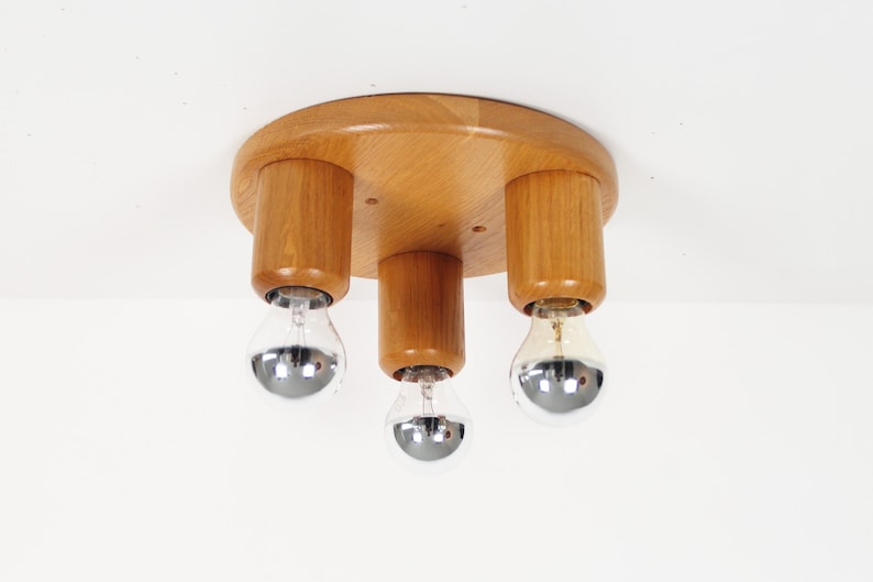 May include: A wooden ceiling light fixture with three cylindrical light holders. The fixture has a round, light-brown wooden base. Each holder has a silver-colored bulb. The light fixture is mounted on a white ceiling.