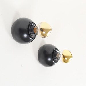 May include: Two black, spherical wall sconces with gold-coloured bulb covers. The sconces have a modern design and are mounted on a white wall. The light fixtures are designed to be mounted on a wall.