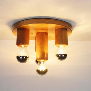 K&ouml;nnte beinhalten: Eine Deckenleuchte aus Holz mit drei Gl&uuml;hbirnen. Die Leuchte hat eine runde Basis und drei zylindrische Arme, die von der Basis nach unten verlaufen. Jeder Arm hat am Ende eine Gl&uuml;hbirne.