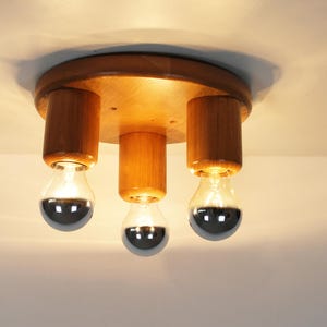 May include: A wooden ceiling light fixture with three exposed bulbs. The fixture has a circular wooden base and cylindrical wooden supports for the bulbs. The bulbs have a silver, reflective coating on the bottom half.