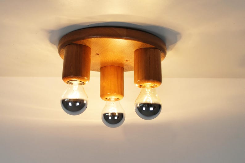 May include: A wooden ceiling light fixture with three exposed bulbs. The fixture has a circular wooden base and cylindrical wooden supports for the bulbs. The bulbs have a silver, reflective coating on the bottom half.