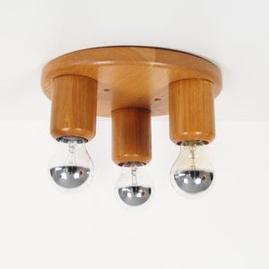 May include: A wooden ceiling light fixture with three light sockets. The fixture has a round, light-colored wooden base with three cylindrical wooden supports. Each support holds a silver-bottomed light bulb.