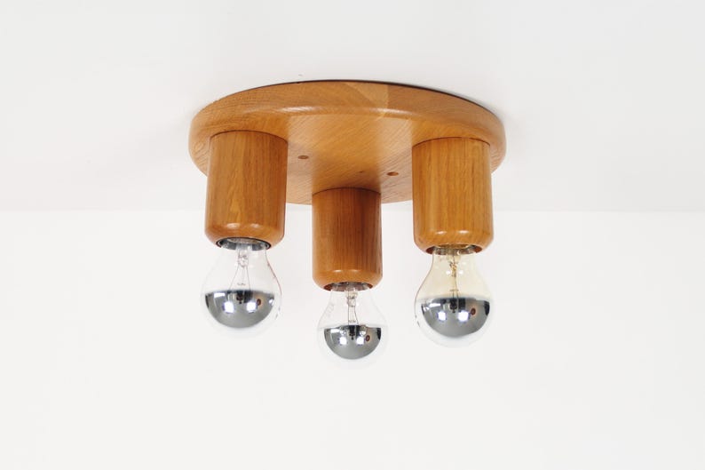 May include: A wooden ceiling light fixture with three light sockets. The fixture has a round, light-colored wooden base with three cylindrical wooden supports. Each support holds a silver-bottomed light bulb.