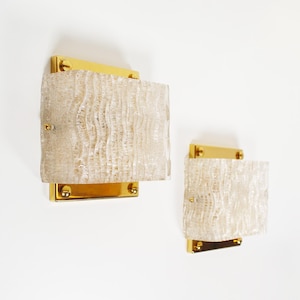May include: Two wall sconces with textured, off-white glass panels mounted on gold-toned metal brackets. The sconces have a rectangular shape and are designed for indoor lighting. The glass has a wavy, organic pattern.