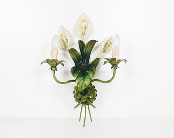 Peut inclure: Applique murale vintage au design floral. L'applique présente des feuilles et des supports de lumière en métal peint en vert et blanc en forme de fleurs. L'applique a trois ampoules et est fixée sur un mur blanc. Le design général rappelle une composition florale stylisée.