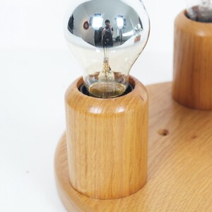 May include: A wooden light fixture with two light bulbs. The light bulbs are silver and clear. The fixture is made of light-colored wood and has a round base. The light bulbs are in cylindrical wooden holders.