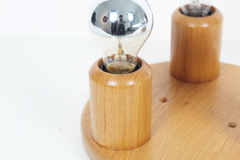 May include: A wooden light fixture with two light bulbs. The light bulbs are silver and clear. The fixture is made of light-colored wood and has a round base. The light bulbs are in cylindrical wooden holders.