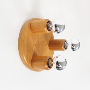 May include: A wooden, round wall sconce with three light fixtures. The sconce has a natural wood finish and chrome-colored, spherical light bulbs. The light fixtures are cylindrical and extend from the wooden base.