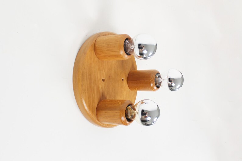 May include: A wooden, round wall sconce with three light fixtures. The sconce has a natural wood finish and chrome-colored, spherical light bulbs. The light fixtures are cylindrical and extend from the wooden base.