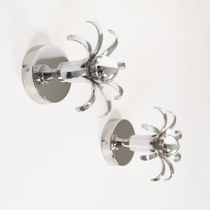 May include: Two chrome wall hooks. Each hook has a circular base and a central stem, with a decorative, flower-like design. The hooks are mounted on a white wall, offering a modern storage solution. Ideal for coats or towels.
