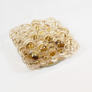 May include: A square flush mount ceiling light fixture featuring a cluster of amber-tinted glass bubbles. The glass elements are arranged in a grid pattern, creating a textured, multi-dimensional surface that reflects light across the translucent spheres.