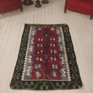 May include: A traditional Turkish kilim rug with a red, blue, green, and white geometric pattern. The rug has a fringed edge and is made of wool.