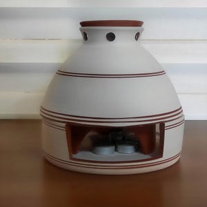 Puede incluir: Un calentador de aceite de cerámica blanca con borde y detalles marrones. El calentador tiene forma redondeada con una abertura rectangular en la base y una serie de rayas marrones. La parte superior tiene varios agujeros pequeños.